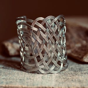Puede incluir: Un brazalete tipo puño de tono plateado con un intrincado diseño de celosía calada, creado a partir de múltiples hilos metálicos finos entrelazados. La estructura forma un patrón continuo de bucles y curvas que se superponen entre sí.