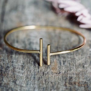 Può includere: Un bracciale aperto color oro caratterizzato da un sottile cerchio che termina con due barre verticali perpendicolari all'apertura centrale. Il metallo presenta una finitura liscia e lucida che riflette la luce lungo la struttura curva.