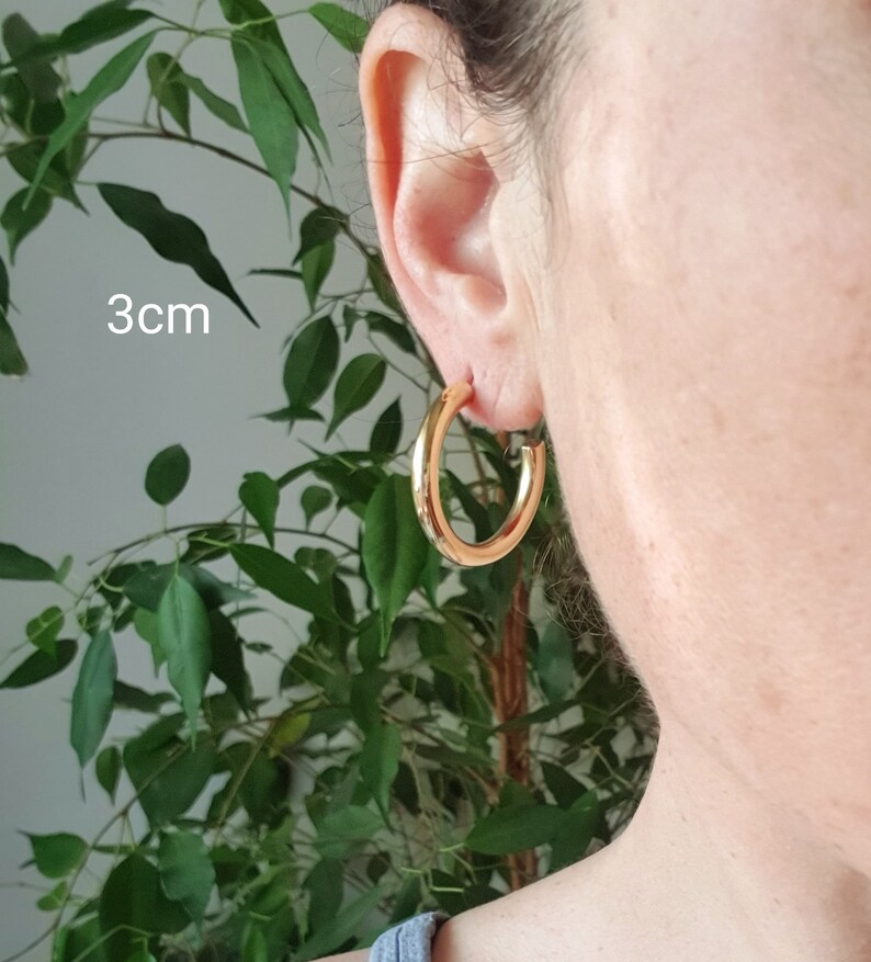 May include: Gold hoop earrings with a diameter of 3 cm. The earrings are shown on a person's ear.