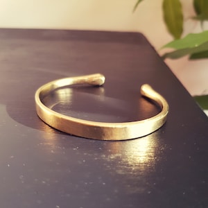 May include: A simple gold-toned bangle bracelet with a smooth, slightly rounded surface. The bracelet is open ended and has a slightly hammered texture.