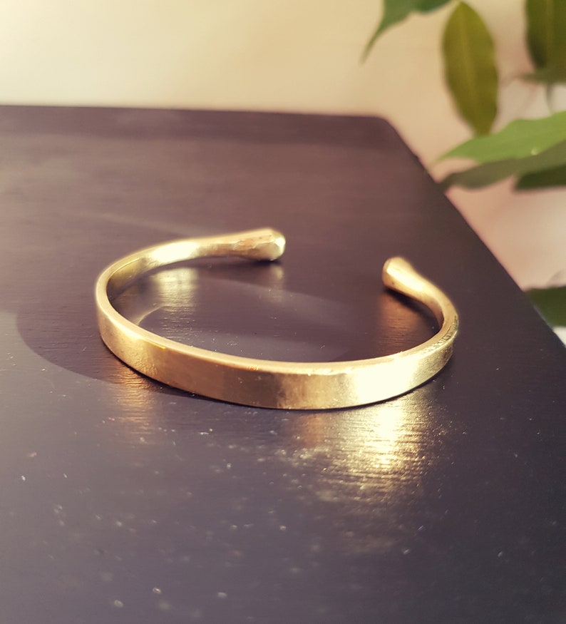 May include: A simple gold-toned bangle bracelet with a smooth, slightly rounded surface. The bracelet is open ended and has a slightly hammered texture.