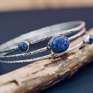 Puede incluir: Una pulsera de plata con tres bandas, con un diseño texturizado. La pulsera está adornada con tres piedras de lapislázuli azules. La joya se exhibe sobre un trozo de madera sobre un fondo oscuro.