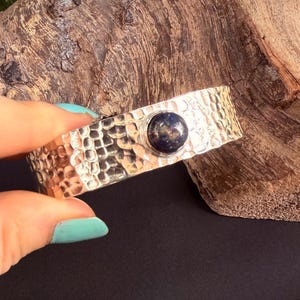 Puede incluir: Una pulsera de puño plateada con una textura martillada y una piedra redonda azul oscuro en el centro. La pulsera se muestra sobre un trozo de madera.