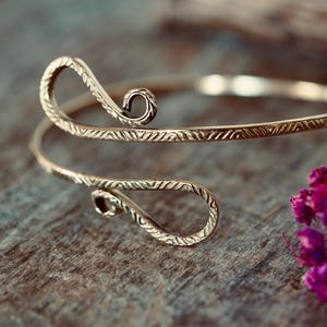 Peut inclure: Un bracelet manchette en métal doré au design ouvert avec deux extrémités en forme de spirale. La surface du bracelet est ornée d'un motif géométrique gravé répétitif qui s'étend sur toute la longueur de la bande métallique.