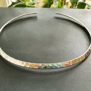 May include: A silver hammered choker necklace. The necklace is made from a single piece of metal that is bent into a circle and then hammered to create a textured surface.