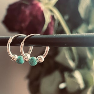May include: Two silver hoop earrings with turquoise beads. The earrings are hanging from a black bar.
