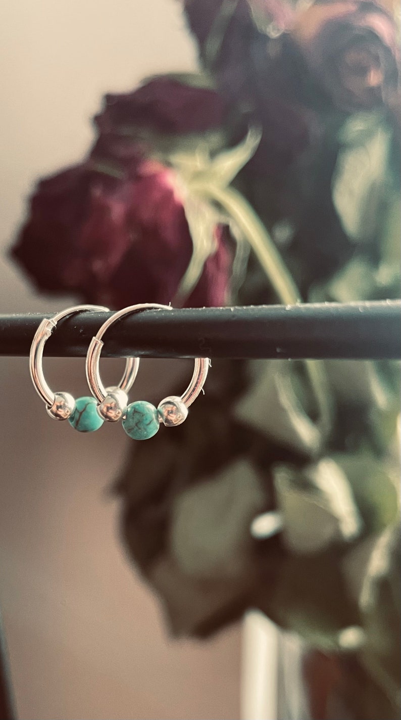 May include: Two silver hoop earrings with turquoise beads. The earrings are hanging from a black bar.