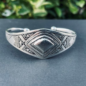 May include: A silver bracelet with a geometric design. The bracelet is made of metal and has a diamond-shaped center piece.