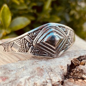 May include: A silver bracelet with a geometric design. The bracelet has a raised diamond shape in the center and is decorated with intricate patterns.