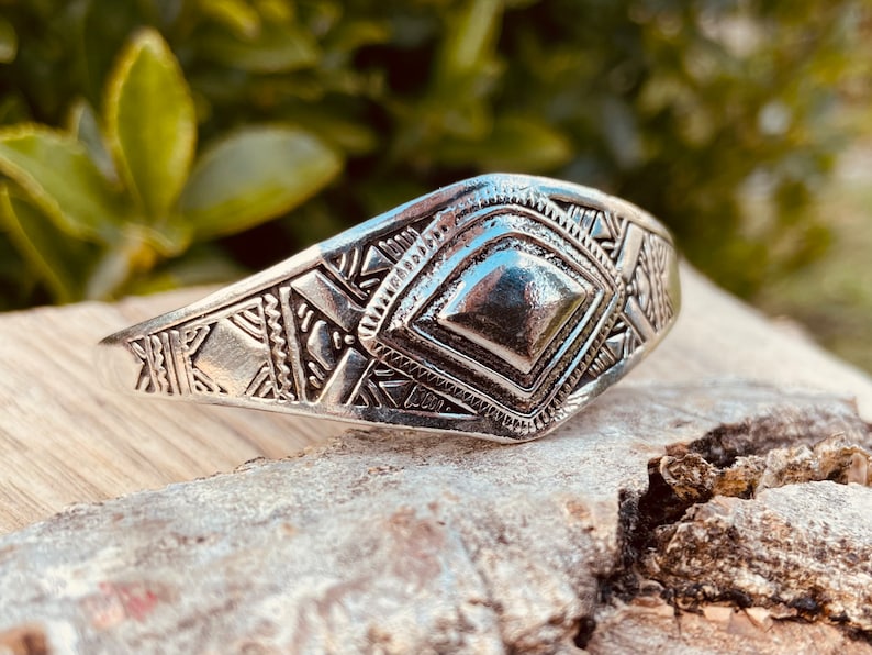 May include: A silver bracelet with a geometric design. The bracelet has a raised diamond shape in the center and is decorated with intricate patterns.