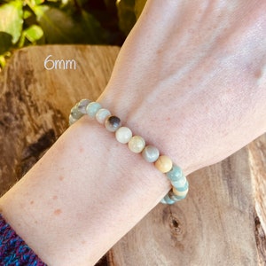 May include: A bracelet made of green, brown, and white 6mm round beads. The bracelet is on a person's wrist.