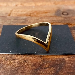 Może przedstawiać: Złoty pierścionek w kształcie litery V. Pierścionek wykonany jest z gładkiego, błyszczącego metalu.