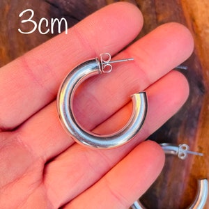 May include: Silver hoop earrings with a 3cm diameter. The earrings are shown on a hand.