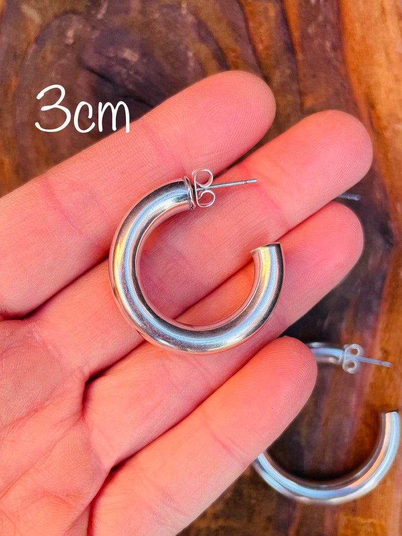 May include: Silver hoop earrings with a 3cm diameter. The earrings are shown on a hand.