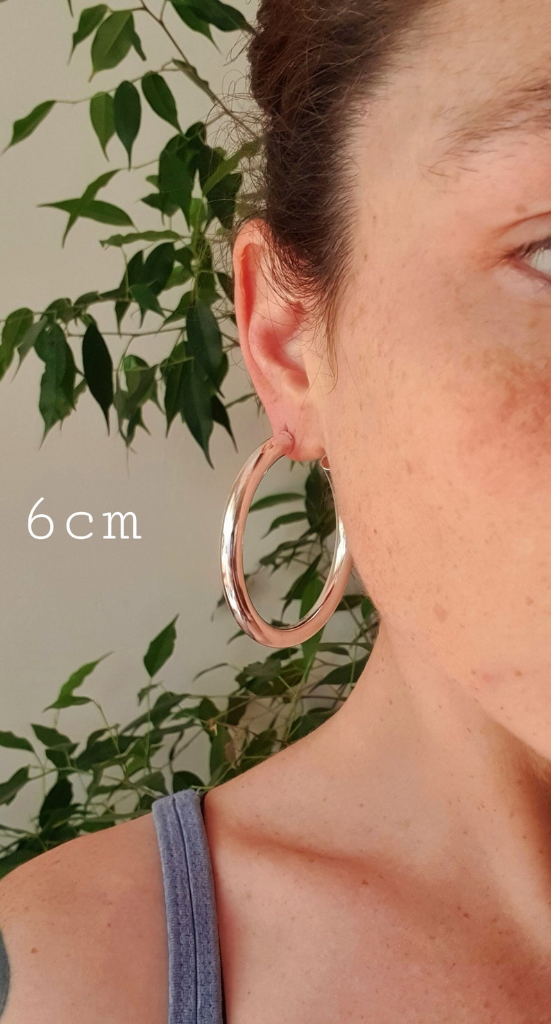 May include: Silver hoop earrings with a diameter of 6 cm. The earrings are shown on a person's ear.