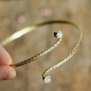 Puede incluir: Un brazalete dorado con un diseño texturizado. El brazalete presenta dos pequeñas gemas redondas y claras en cada extremo. El brazalete es sostenido por una mano, mostrando su elegante diseño abierto.