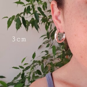 May include: Silver hoop earrings, 3 cm in diameter. The earrings are shown on a person's ear.