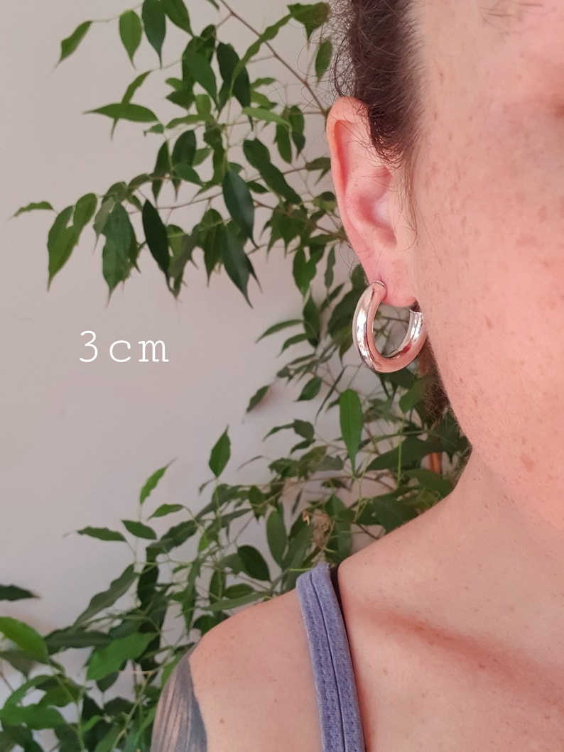 May include: Silver hoop earrings, 3 cm in diameter. The earrings are shown on a person's ear.