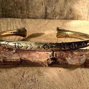 May include: A gold-toned metal bracelet with an intricate etched design. The bracelet is open and has a simple, rounded shape.