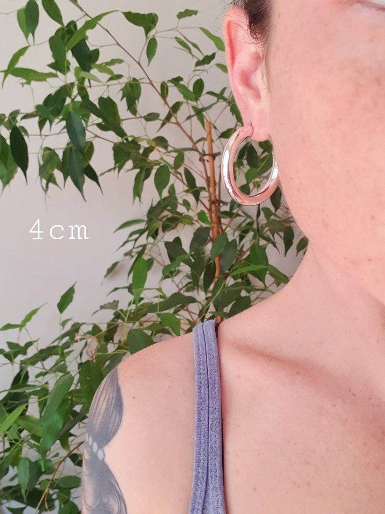 May include: Silver hoop earrings with a diameter of 4 cm. The earrings are shown on a person's ear.
