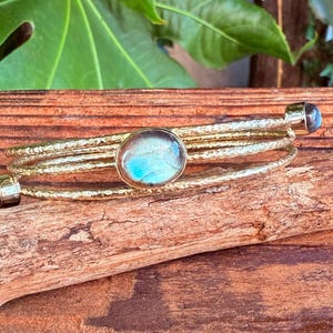 Könnte beinhalten: Ein goldfarbenes Armband mit strukturiertem Design, mit drei Bändern und drei runden, hellblauen Edelsteinakzenten. Das Armband liegt auf einem Holzstück vor einem hölzernen Hintergrund.