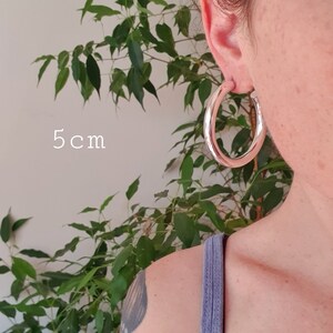 May include: Silver hoop earrings with a diameter of 5 cm. The earrings are shown on a person's ear.