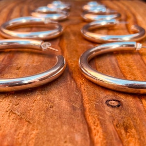 May include: A set of six silver hoop earrings, arranged in a row on a wooden surface. The hoops are thick and have a smooth, polished finish.