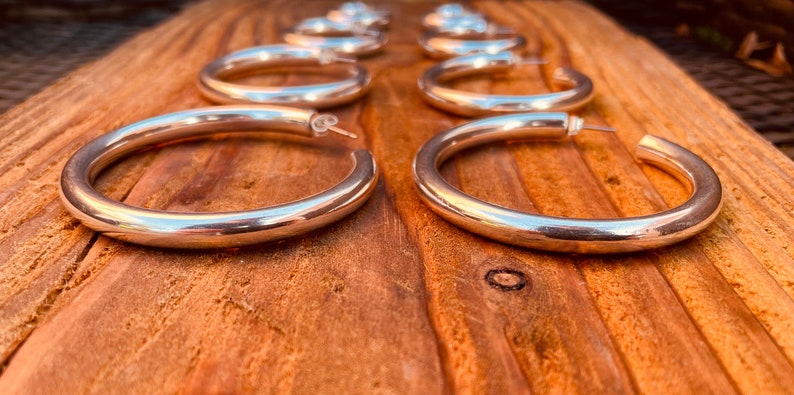 May include: A set of six silver hoop earrings, arranged in a row on a wooden surface. The hoops are thick and have a smooth, polished finish.
