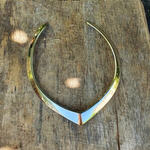 May include: A silver metal necklace with a V-shaped design. The necklace is lying on a wooden surface.