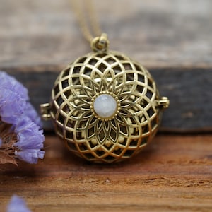 May include: A gold-toned spherical locket pendant featuring an intricate openwork geometric lattice pattern and a small, round, white cabochon stone at the center. The pendant hangs from a thin gold-toned chain.