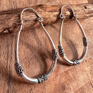 May include: A pair of silver-colored hoop earrings with an elongated oval shape. The earrings have decorative bands with an intricate design. The earrings are displayed on a piece of wood.