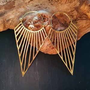 Peut inclure: Une paire de boucles d'oreilles créoles géométriques dorées avec un motif en forme de soleil. Les boucles d'oreilles sont en métal et ont une forme triangulaire.