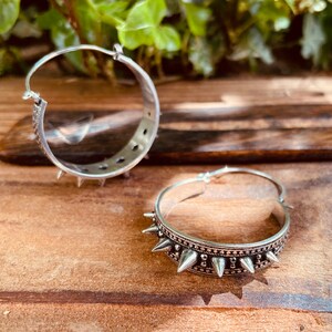 May include: A pair of silver hoop earrings with a row of spikes around the edge. The earrings are on a wooden surface.