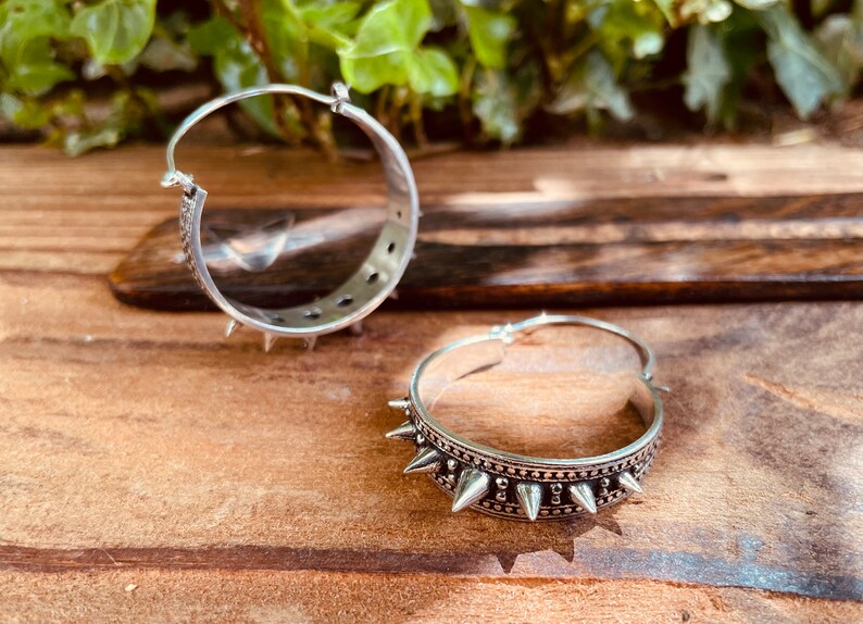 May include: A pair of silver hoop earrings with a row of spikes around the edge. The earrings are on a wooden surface.