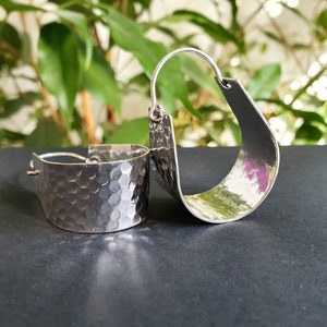 May include: A pair of silver hoop earrings with a hammered texture. The earrings are curved and have a slightly oval shape.