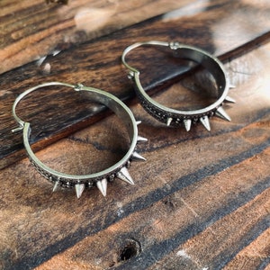 May include: A pair of silver hoop earrings with a row of spikes around the edge. The earrings are on a wooden surface.