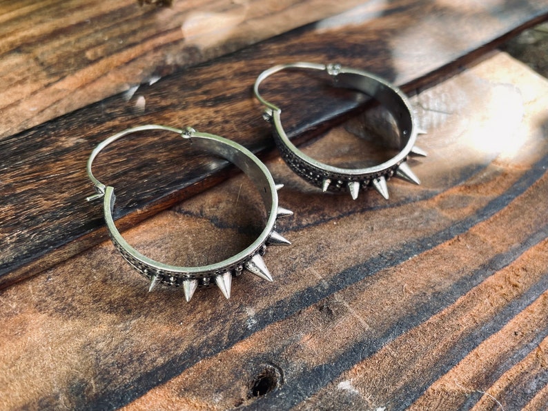 May include: A pair of silver hoop earrings with a row of spikes around the edge. The earrings are on a wooden surface.