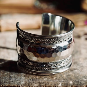 Peut inclure: Un large bracelet manchette argenté présentant une bande centrale à texture martelée, encadrée par deux bordures décoratives aux motifs complexes en forme de corde et de zigzag. Le métal a une finition polie et réfléchissante.