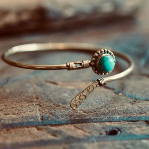 Op de afbeelding: Een goudkleurige open armband met een ronde, helder turquoise steen in een geschulpte zetting aan het ene uiteinde en een plat, getextureerd, bladachtig metalen detail aan het andere uiteinde, verbonden door een dunne, gladde metalen band.
