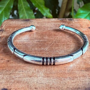 May include: A silver cuff bracelet with a rounded design. The bracelet features black vertical lines and geometric shapes. The ends of the bracelet have a rounded ball shape. The bracelet is displayed on a wooden surface.