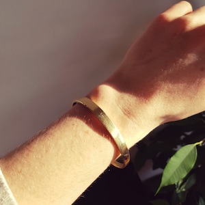 May include: A gold-toned metal bracelet on a person's wrist. The bracelet is simple and has a smooth, polished finish.