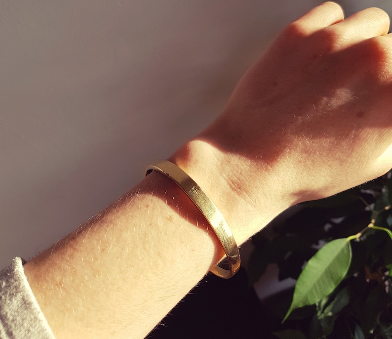 May include: A gold-toned metal bracelet on a person's wrist. The bracelet is simple and has a smooth, polished finish.