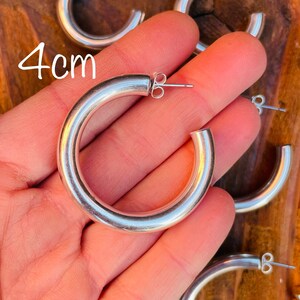 May include: Silver hoop earrings with a 4 cm diameter. The earrings are shown in a hand, with the back of the earrings visible.