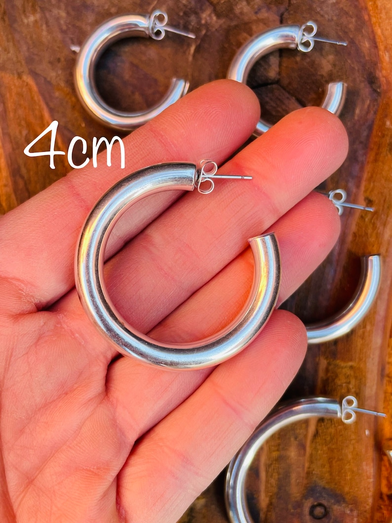 May include: Silver hoop earrings with a 4 cm diameter. The earrings are shown in a hand, with the back of the earrings visible.