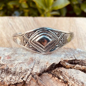 May include: A silver bracelet with a geometric design. The bracelet is made of metal and has a diamond-shaped center piece.
