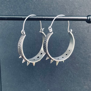 May include: A pair of silver hoop earrings with a textured design and pointed edges. The earrings are hanging on a black stand.