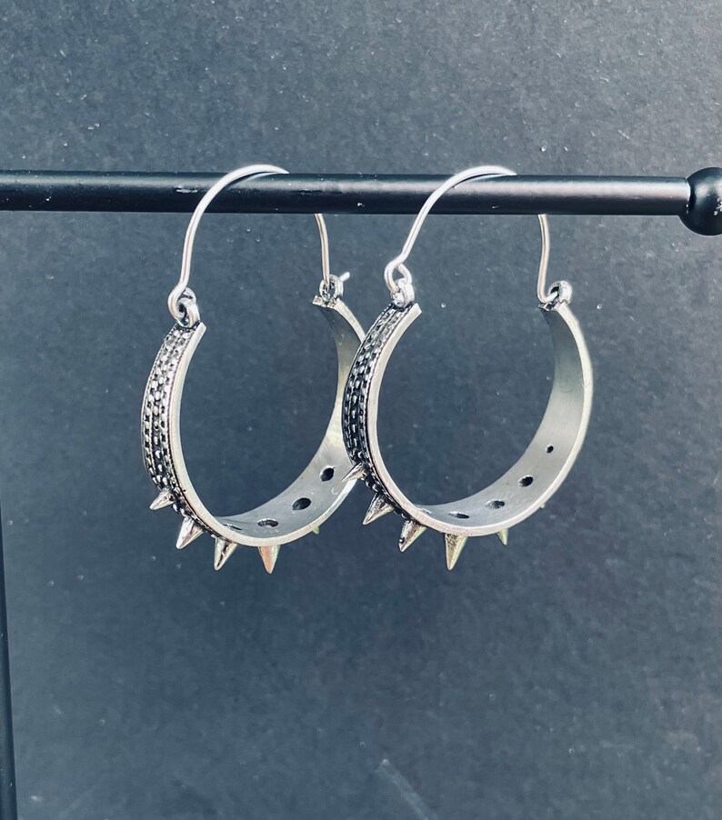 May include: A pair of silver hoop earrings with a textured design and pointed edges. The earrings are hanging on a black stand.