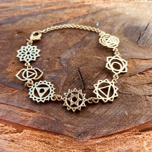Op de afbeelding: Gouden armband met zeven bedels, elk met een ander chakra-symbool. De symbolen zijn ingewikkeld en gedetailleerd, en de armband is gemaakt van een delicate ketting.