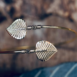 May include: A gold-colored cuff bracelet featuring two leaf-shaped accents. The leaves have detailed veining and are connected to the bracelet's band. The bracelet is a simple, elegant design.