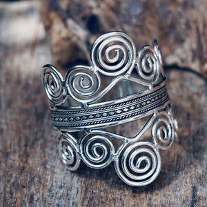 Può includere: Un anello argentato dal design aperto con motivi a spirale. L'anello ha una fascia larga con una sezione centrale dettagliata e strutturata. L'anello è esposto su una superficie di legno, mostrando l'intricato lavoro del metallo.